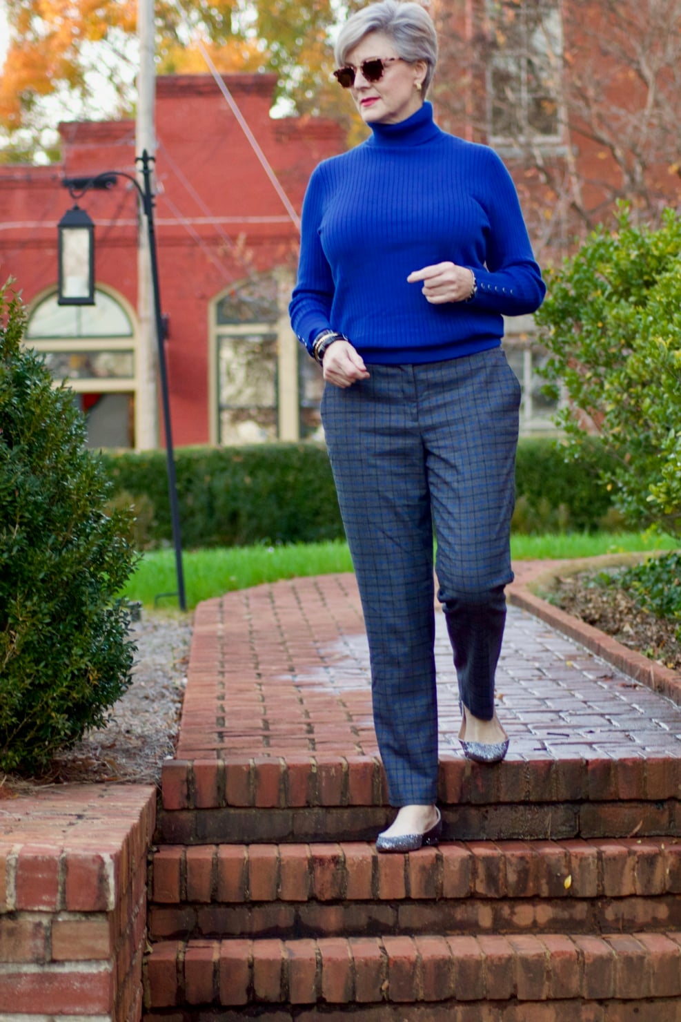 beth from style at a certain age wears plaid pants, cobalt blue turtleneck, green wool coat, blackwatch plaid cashmere scarf, and green beanie
