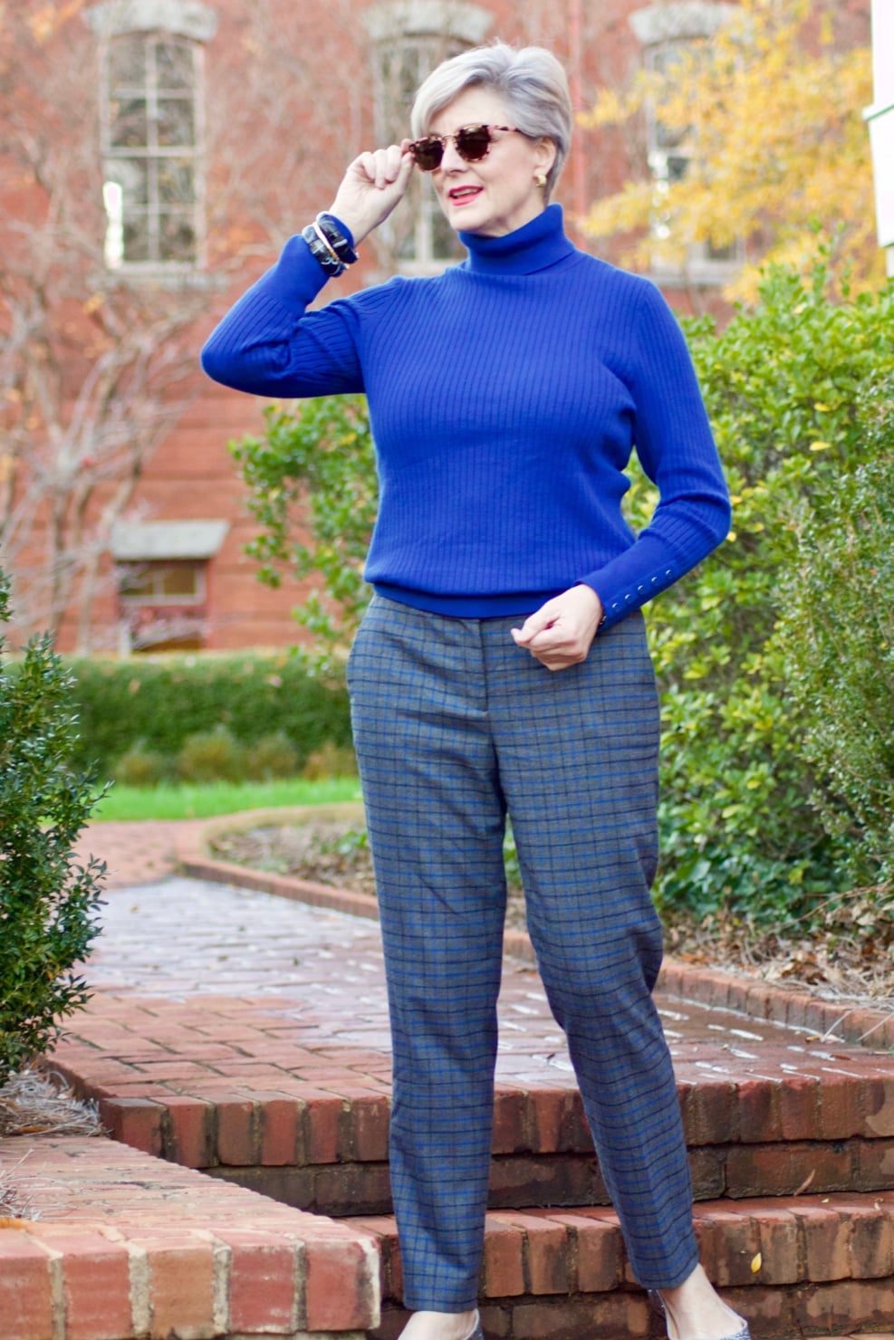 beth from style at a certain age wears plaid pants, cobalt blue turtleneck, green wool coat, blackwatch plaid cashmere scarf, and green beanie