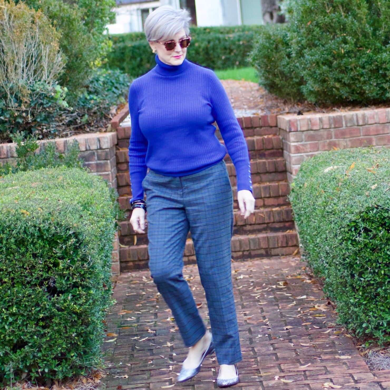 beth from style at a certain age wears plaid pants, cobalt blue turtleneck, green wool coat, blackwatch plaid cashmere scarf, and green beanie