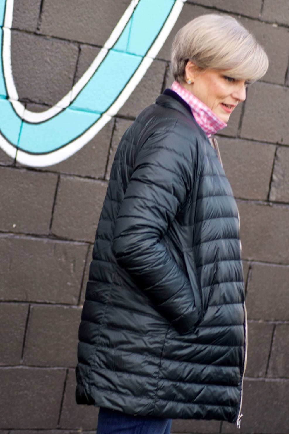 beth from Style at a Certain Age wears a puffer jacket, navy blue knit blazer, novelty sweater, pink gingham check shirt and blue jeans