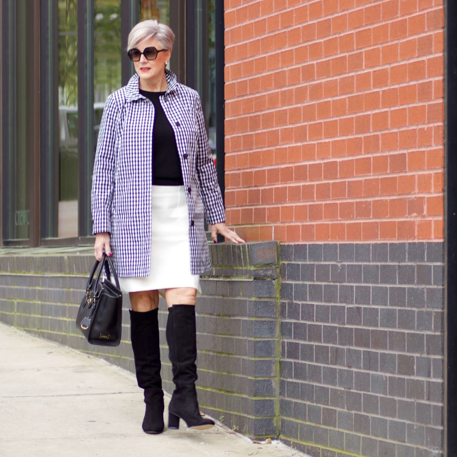 beth from Style at a Certain Age wears a gingham raincoat, white skirt, black sweater, and black boots