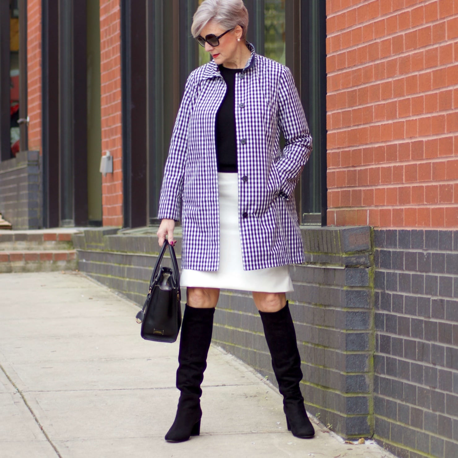 beth from Style at a Certain Age wears a gingham raincoat, white skirt, black sweater, and black boots