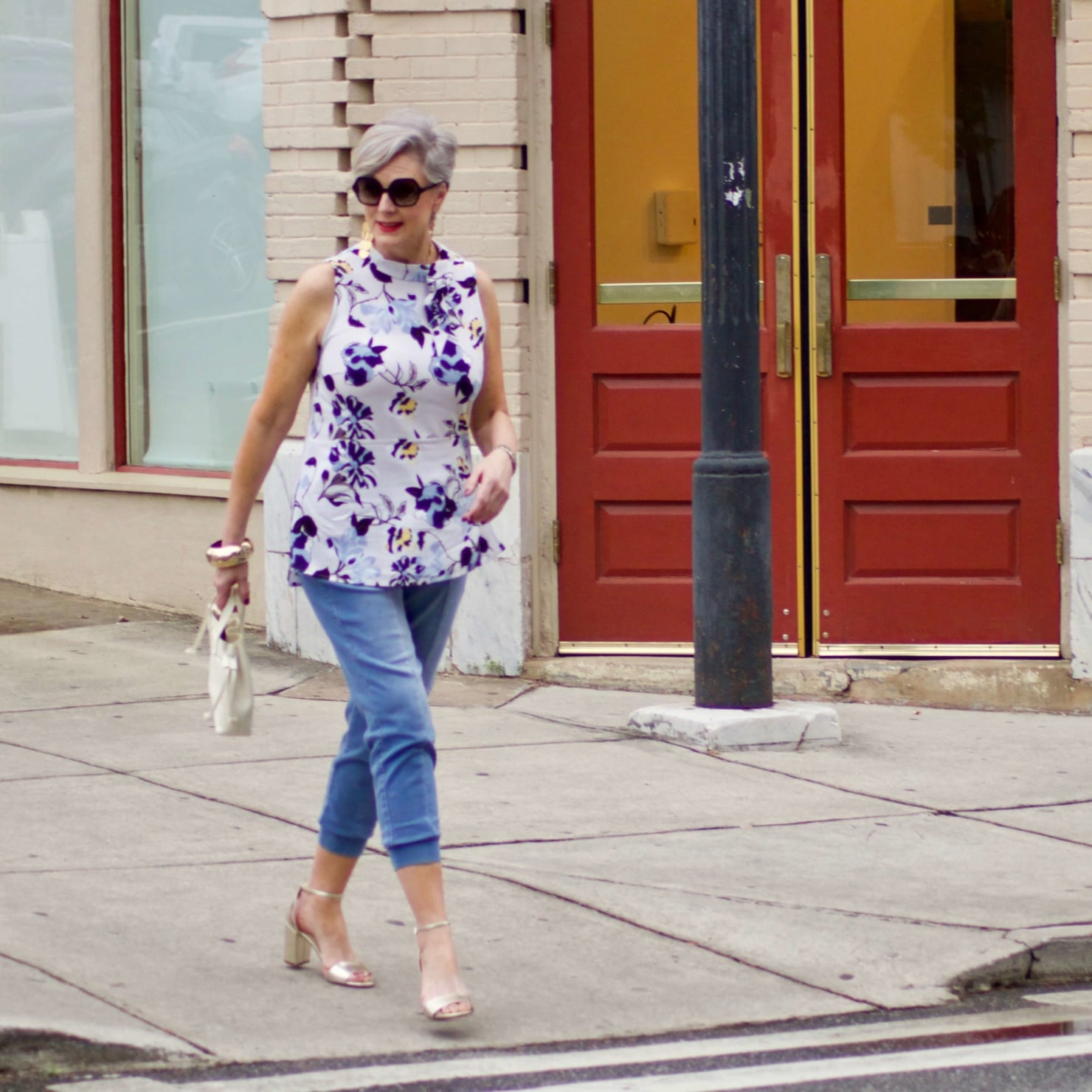 beth from Style at a Certain Age wears a floral top, denim joggers, gold metallic block sandals, and carries a drawstring crossbody handbag. Walmart we dress America