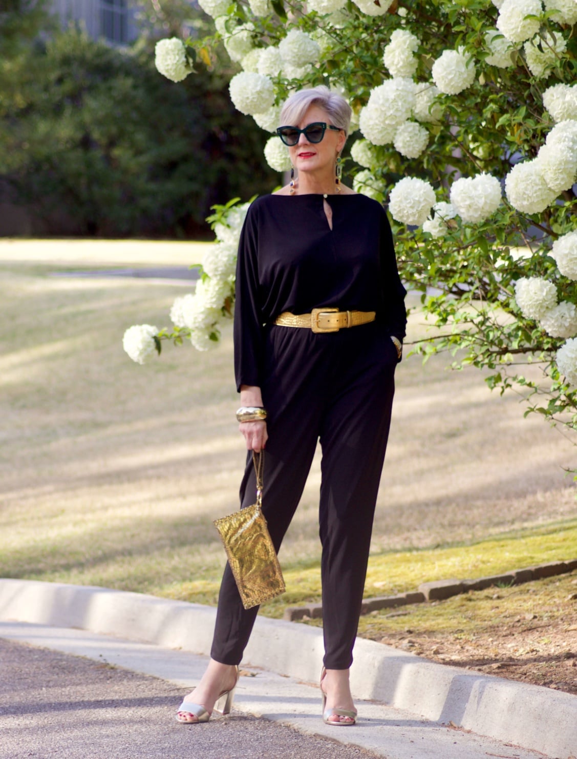 beth from Style at a Certain Age wears a black jumpsuit, gold sandals, gold belt, and gold clutch handbag