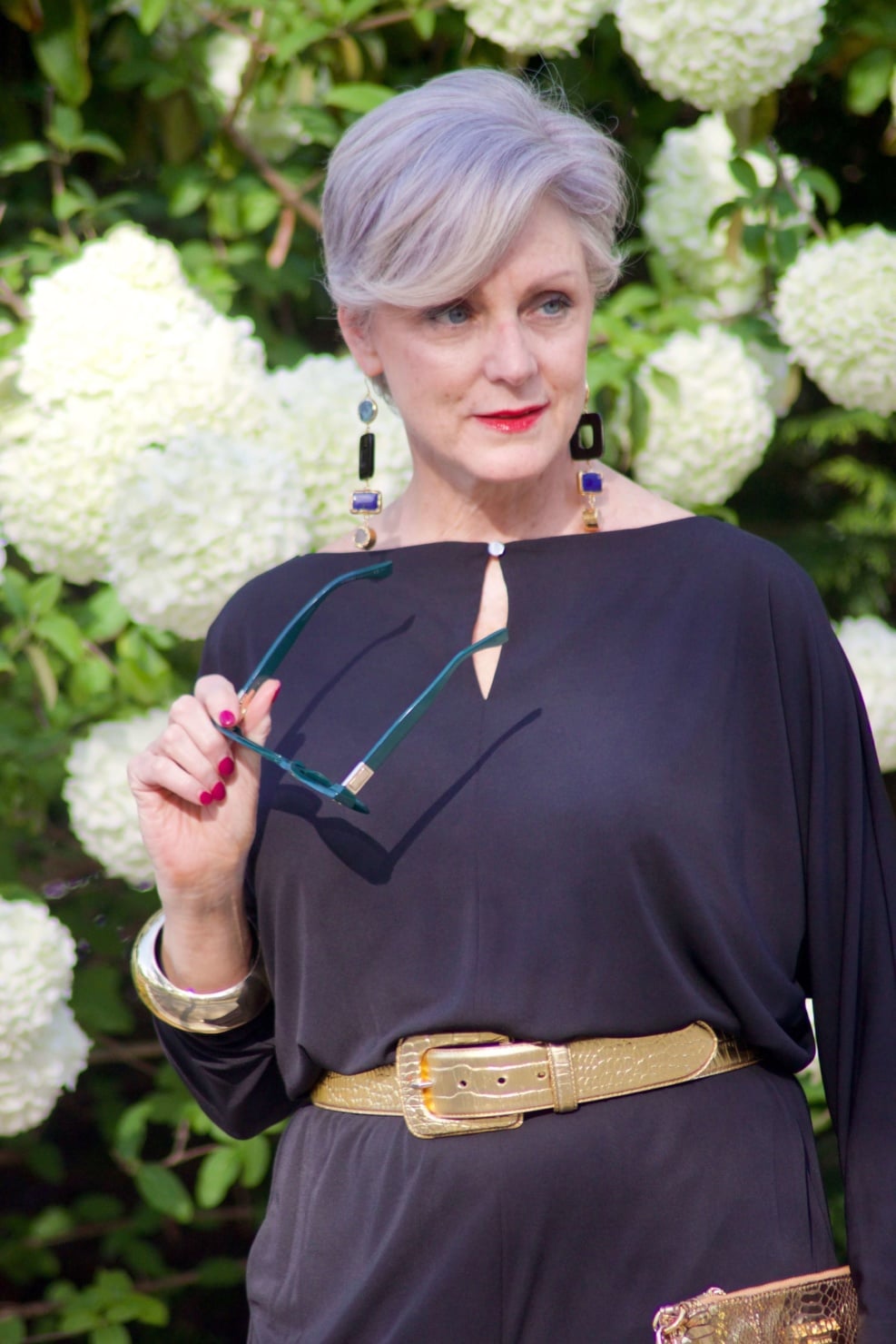 beth from Style at a Certain Age wears a black jumpsuit, gold sandals, gold belt, and gold clutch handbag