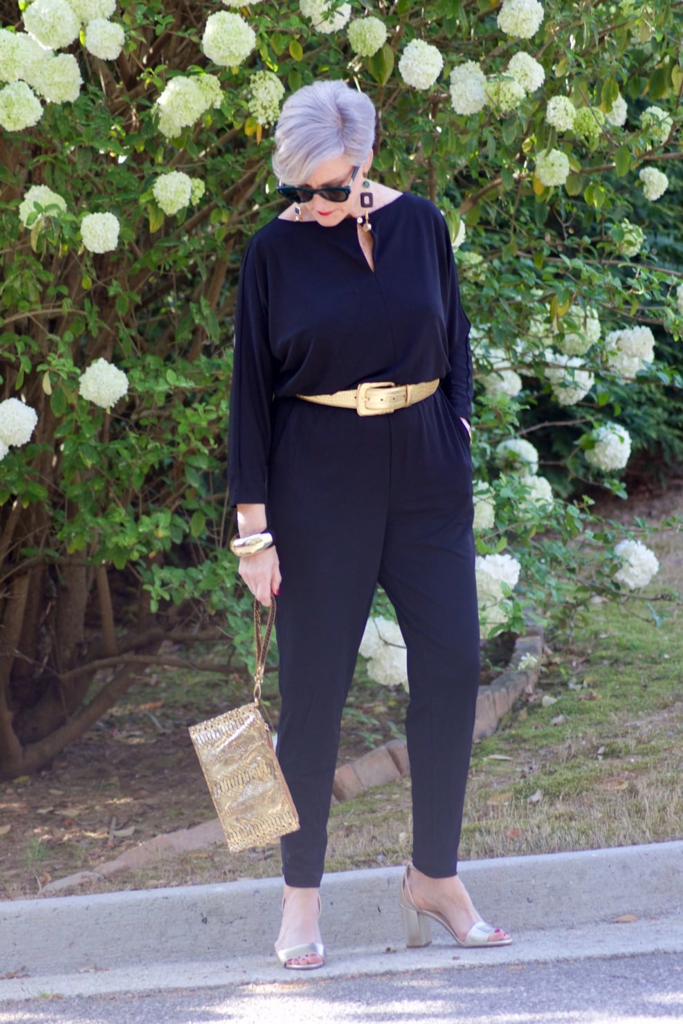 beth from Style at a Certain Age wears a black jumpsuit, gold sandals, gold belt, and gold clutch handbag