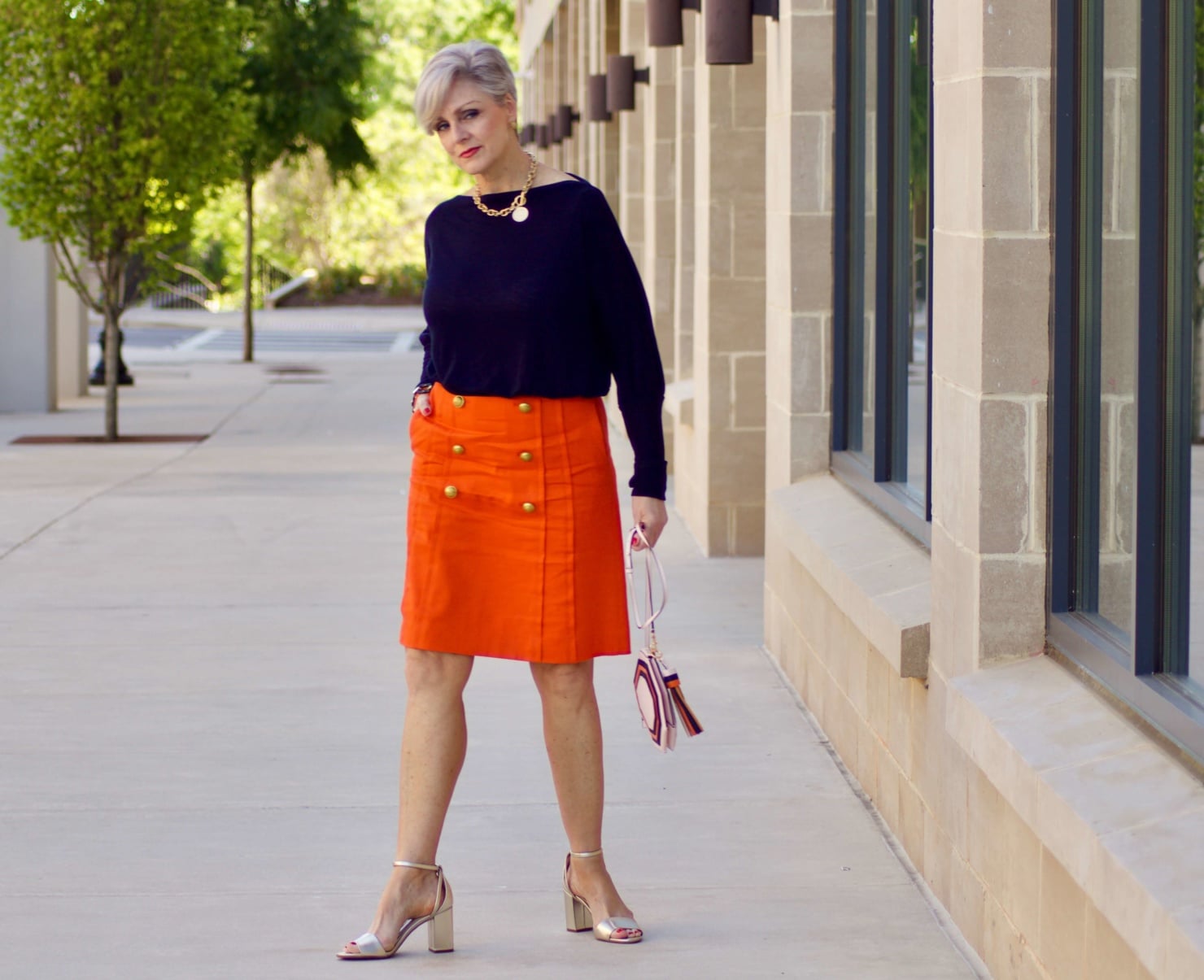 beth from Style at a Certain Age wears a J.Crew orange skirt, blue tee, gold sandals, and Tory Burch mini bag.