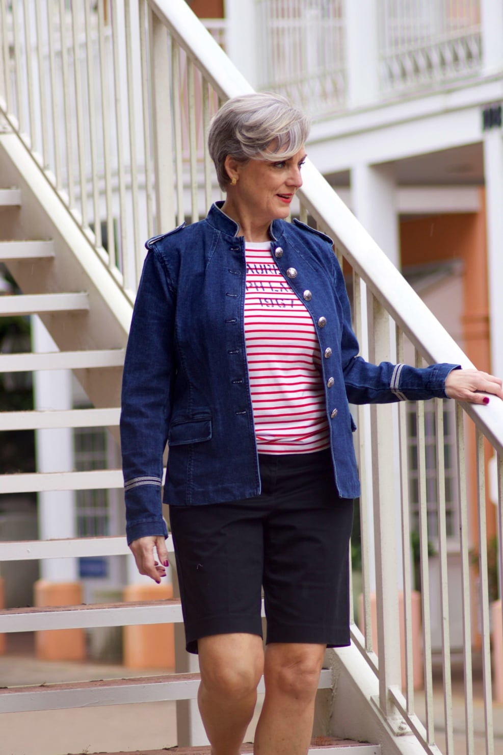 beth from Style at a Certain Age wears a military inspired denim jacket, red stripe tee, black bermuda shorts, and white tennis shoes