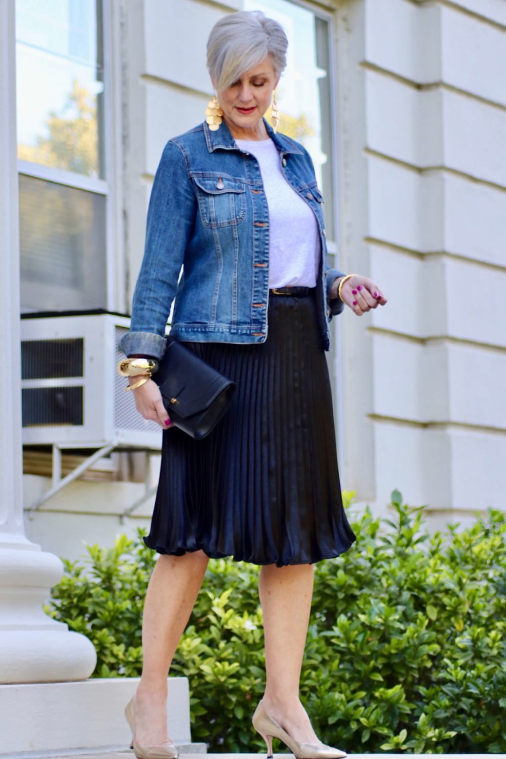 beth from Style at a Certain Age wears a black pleated skirt, white tee, denim jacket, and gold metallic pumps