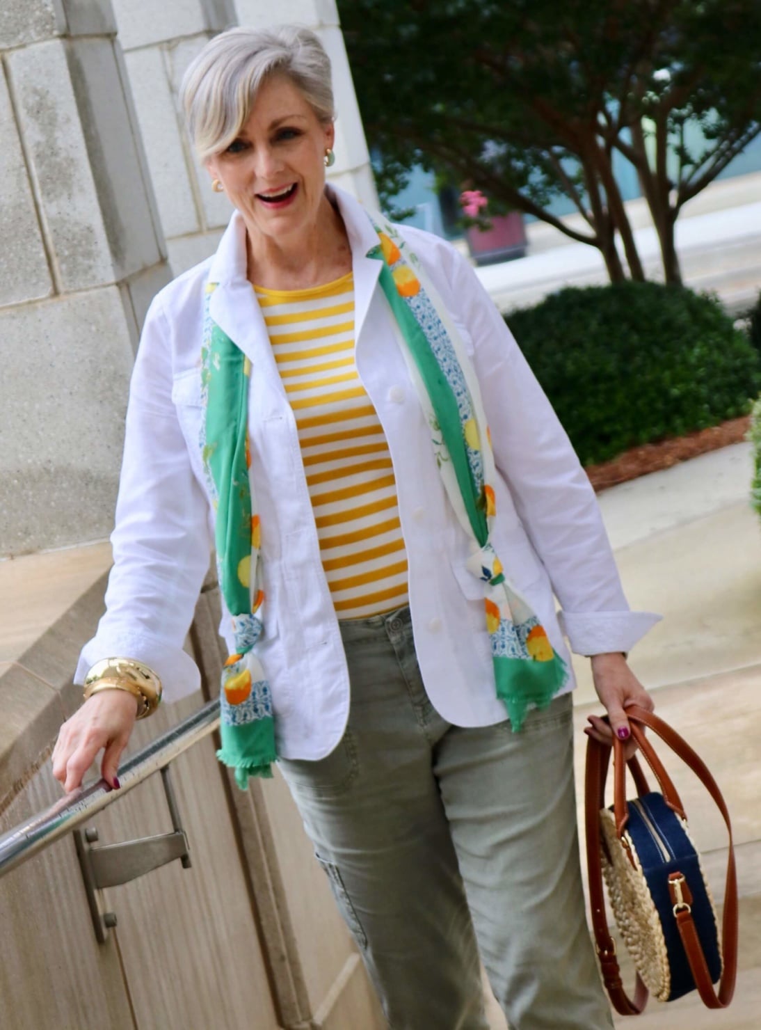 how to wear a jacket for spring and summer beth from Style at a Certain Age wears a white utility jacket, striped tee, green utility pants, yellow sandals, lemon scarf, and cornhusk handbag
