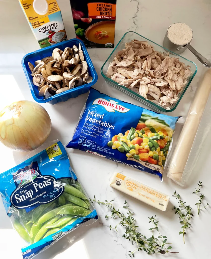 Flat lay of spring chicken pot pie ingredients including heavy whipping cream, chicken broth, sliced mushrooms, shredded rotisserie chicken, all-purpose flour, yellow onion, Birds Eye frozen mixed vegetables, sugar snap peas, butter, fresh thyme, and refrigerated pie crust on a marble counter