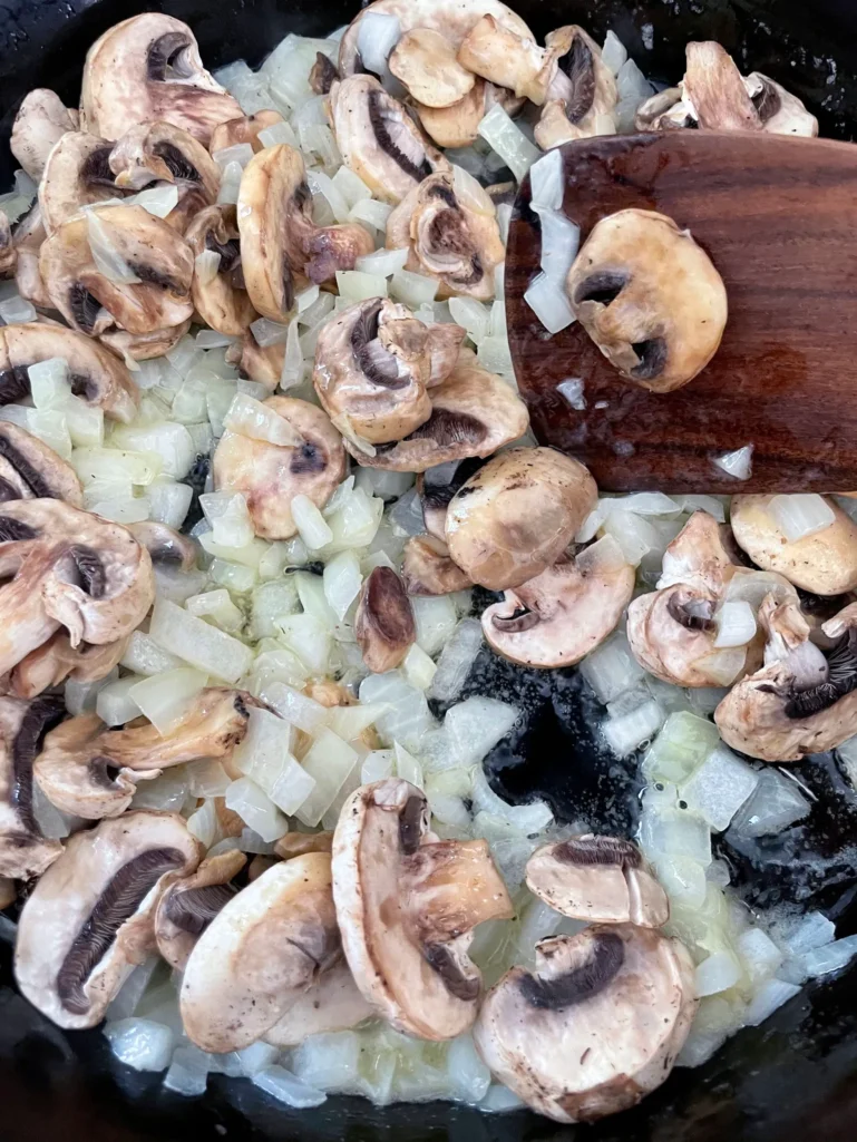 Sliced cremini mushrooms and diced yellow onions sautéing together in a large cast iron skillet with a wooden spoon