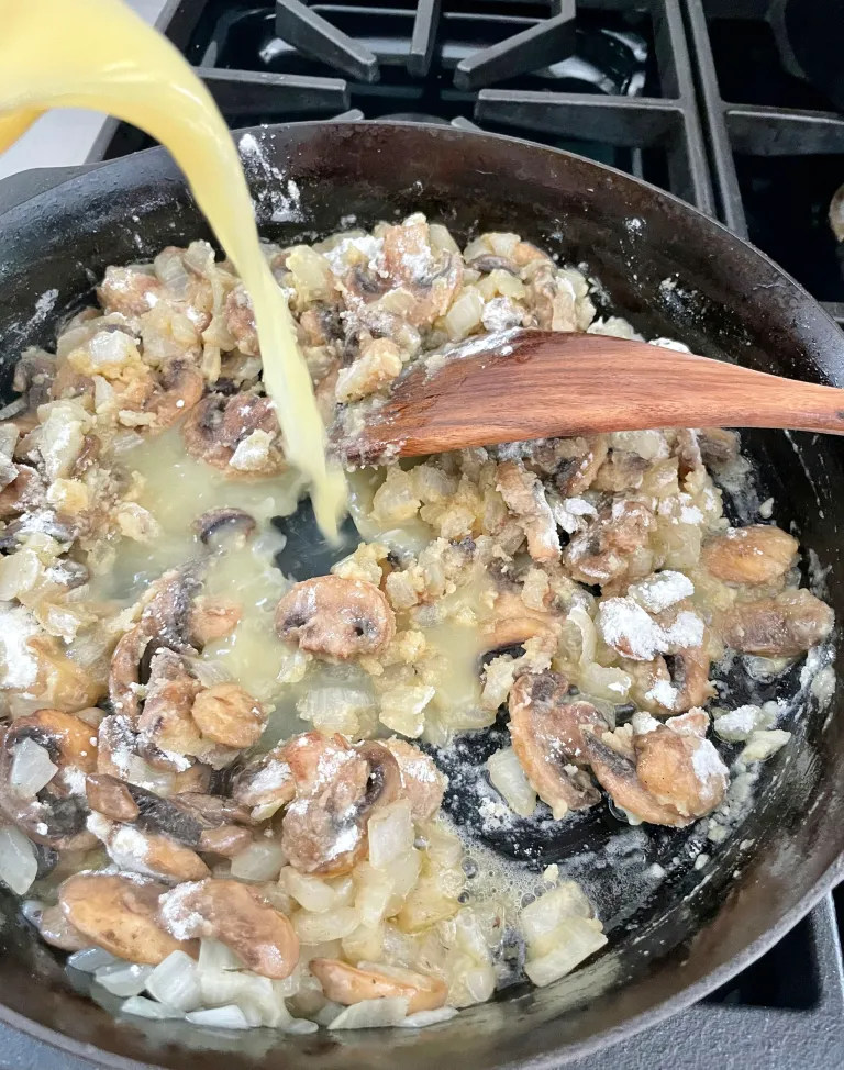 Chicken broth being poured from a yellow pitcher into the flour-coated mushroom and onion mixture in a cast iron skillet