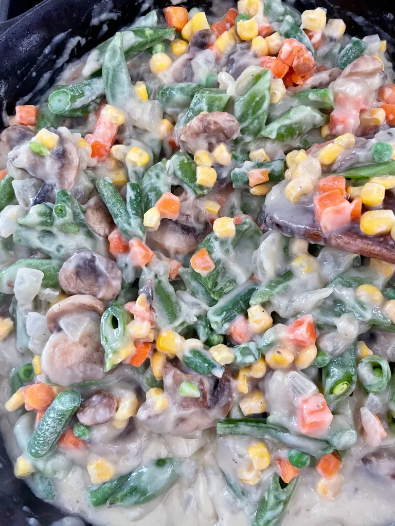 Frozen mixed vegetables including green beans, corn, carrots, and peas stirred into the creamy mushroom sauce in a cast iron skillet