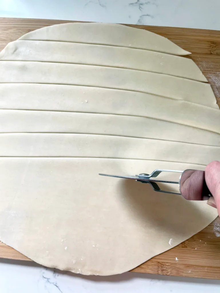 Refrigerated pie dough being cut into even strips with a pastry wheel cutter on a wooden cutting board