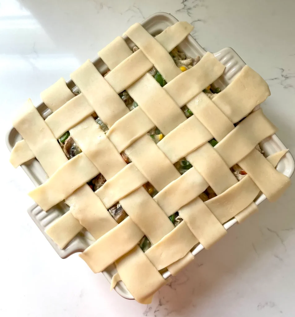 Overhead view of a raw woven lattice pie crust laid over spring chicken pot pie filling in a white square casserole dish