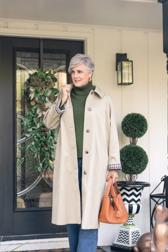Woman wearing trench coat layered over green turtleneck with orange handbag.