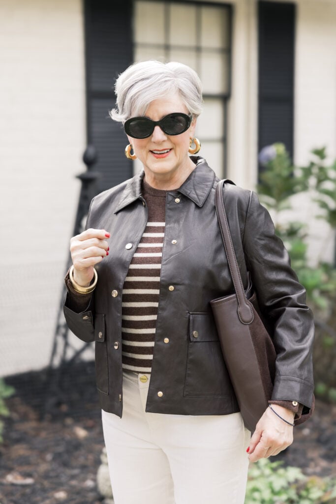 Beth Djalali walking confidently on Tybee Island in a brown faux leather jacket, striped sweater, ivory jeans, and brown suede flats, holding a matching hobo bag in front of a white-brick cottage.