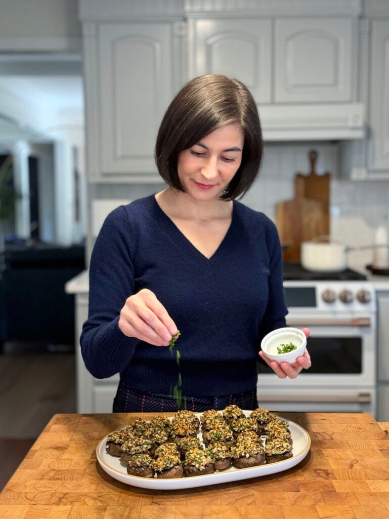 Woman with dark bob haircut wearing navy cashmere sweater garnishing golden-brown stuffed mushrooms with fresh herbs in a bright white kitchen