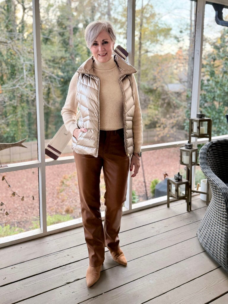 Woman with short silver-blonde hair wearing gold metallic quilted puffer vest over camel ribbed turtleneck sweater, brown faux leather straight-leg pants, and tan suede booties, standing on screened porch