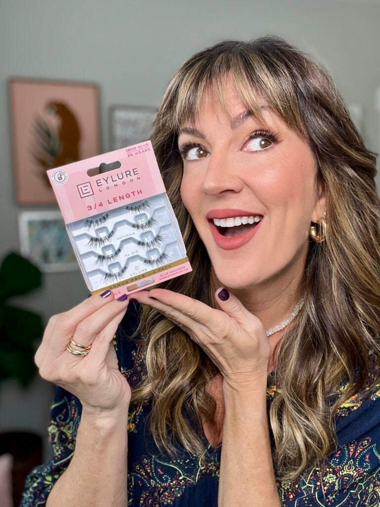 Woman with highlighted brown hair wearing navy floral top holding Eylure false lashes package, smiling at camera with natural makeup