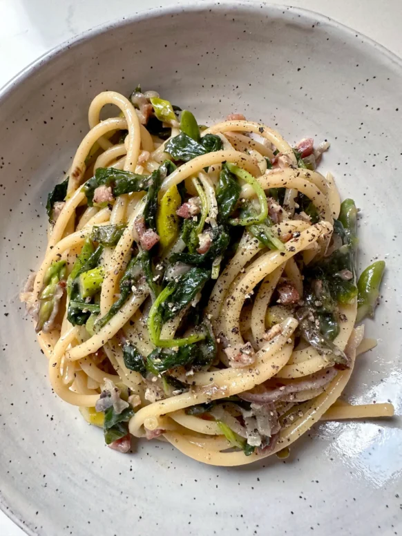 Plated blue cheese bucatini with greens and pancetta in speckled bowl topped with black pepper