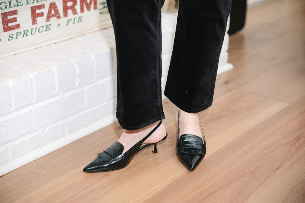Woman over 50 wearing red and black plaid tie-neck blouse with black velvet jeans and black slingback heels standing by decorated fireplace mantel holding glass of red wine