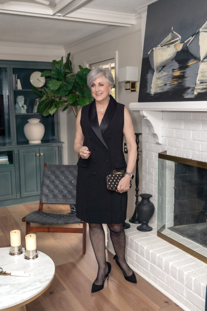Woman over 50 wearing sleeveless black tuxedo dress with satin lapels, patterned black tights, black pumps, and gold polka dot clutch