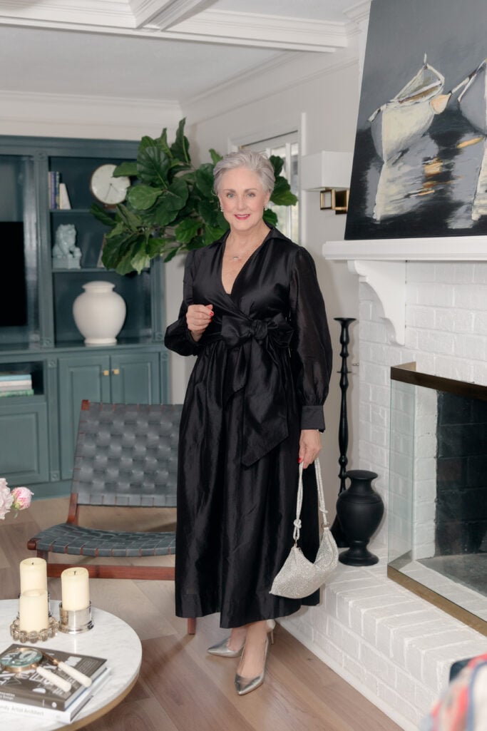 Woman over 50 wearing black taffeta wrap dress with balloon sleeves, bow waist detail, silver metallic pumps, and crystal-embellished evening bag