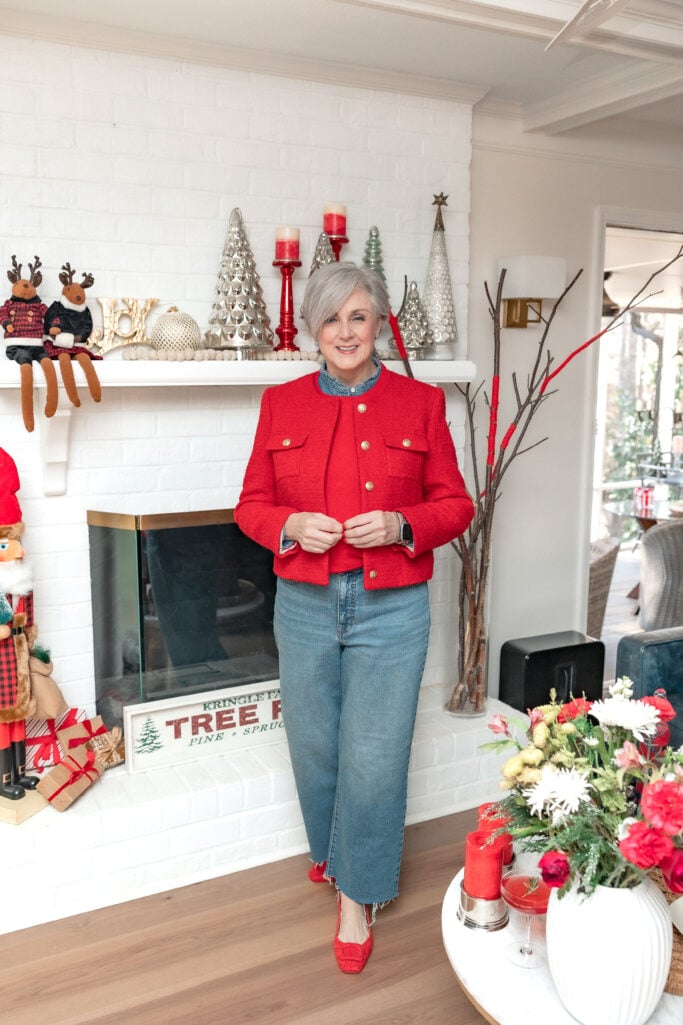 Full-length photo of woman in red holiday jacket with gold button details, light denim jeans, and red flats standing in front of white brick fireplace with Christmas tree farm sign and holiday decorations