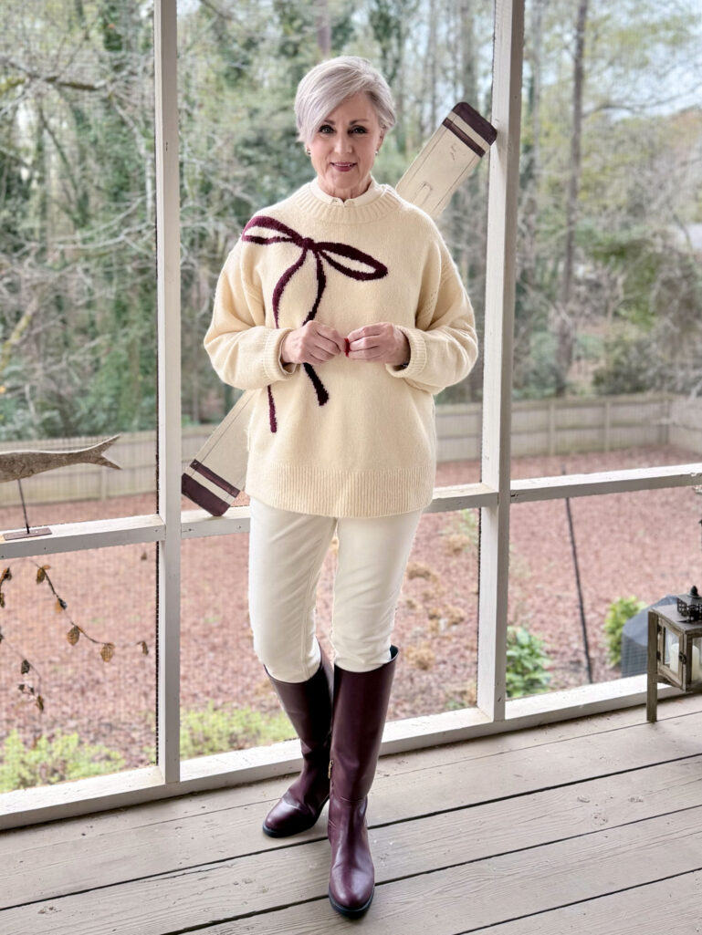 Beth wearing a cream bow-print sweater with burgundy ribbon detail, ivory velveteen pants, and burgundy riding boots on a screened porch