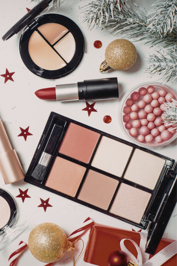 Flat lay of holiday-themed makeup essentials, including a red lipstick, concealer palette, blush and highlighter palette, and pink illuminating powder pearls, styled with gold ornaments, candy canes, red stars, and frosted greenery.