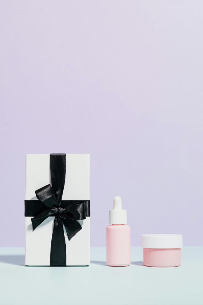 Minimal skincare display featuring a white gift box with black ribbon, a pink serum dropper bottle, and a matching pink cream jar on a soft pastel background.