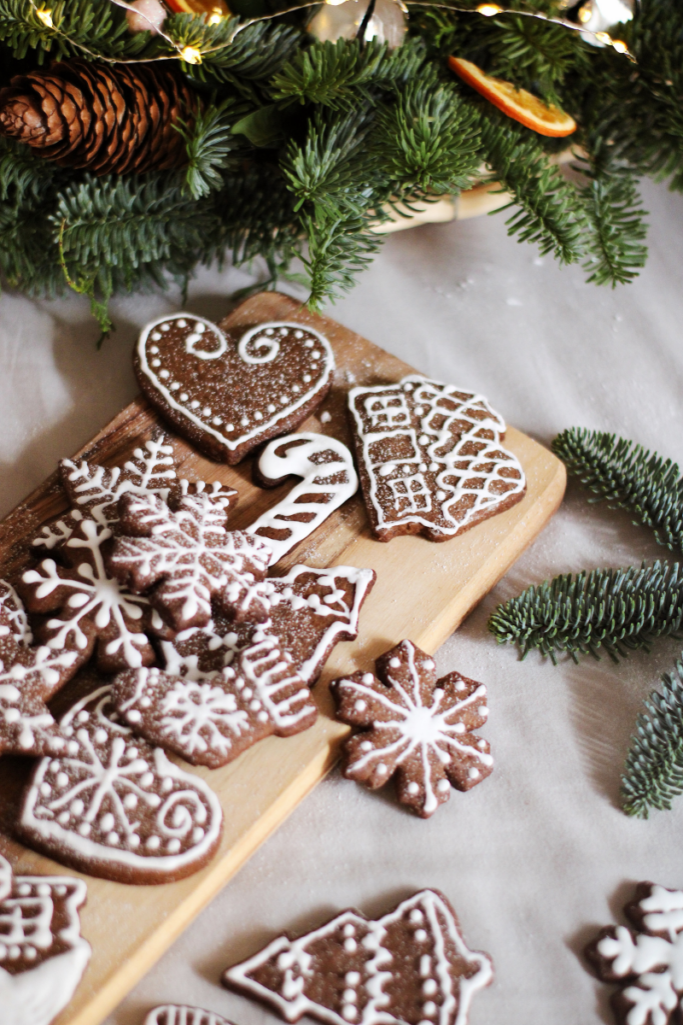 Holiday baking scene with iced gingerbread cookies on a wooden board, surrounded by fresh evergreen branches, pinecones, and warm string lights.