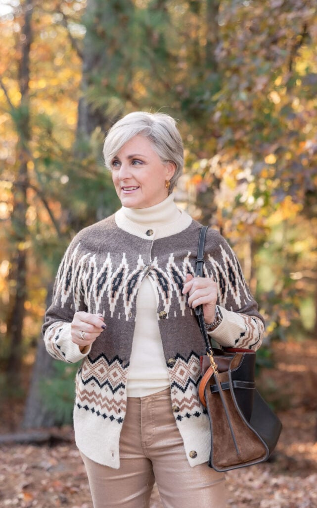 Beth wearing brown and cream fair isle cardigan with dramatic yoke pattern over cream turtleneck with camel pants, black and cognac leather tote, standing on tree-lined path with autumn leaves