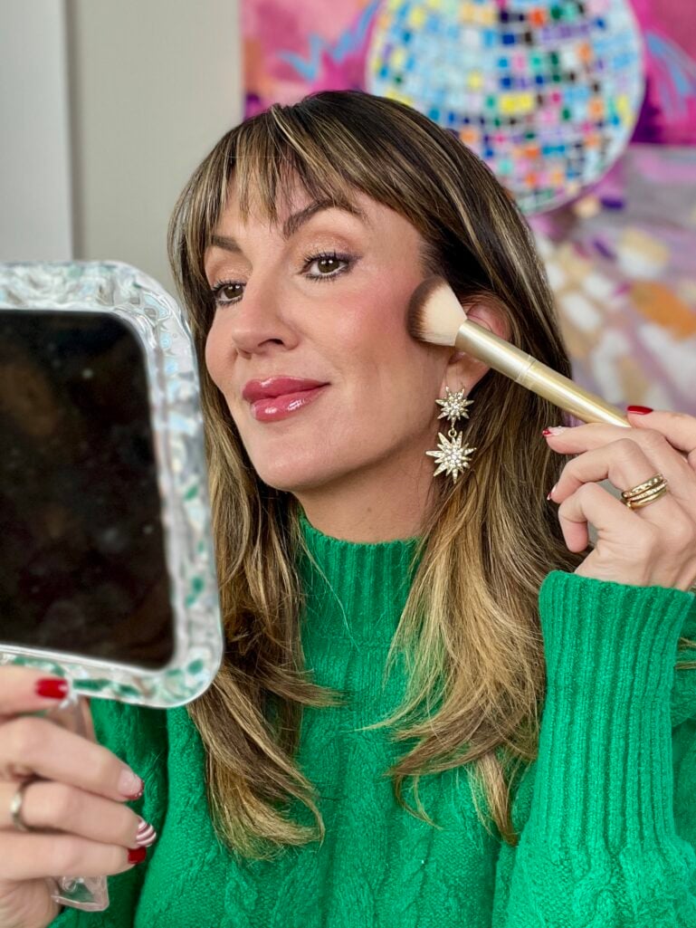 Woman with highlighted brown hair and bangs wearing emerald green cable knit turtleneck sweater, applying blush with gold makeup brush while holding iridescent handheld mirror, wearing gold celestial star earrings and festive red and white candy cane nail art