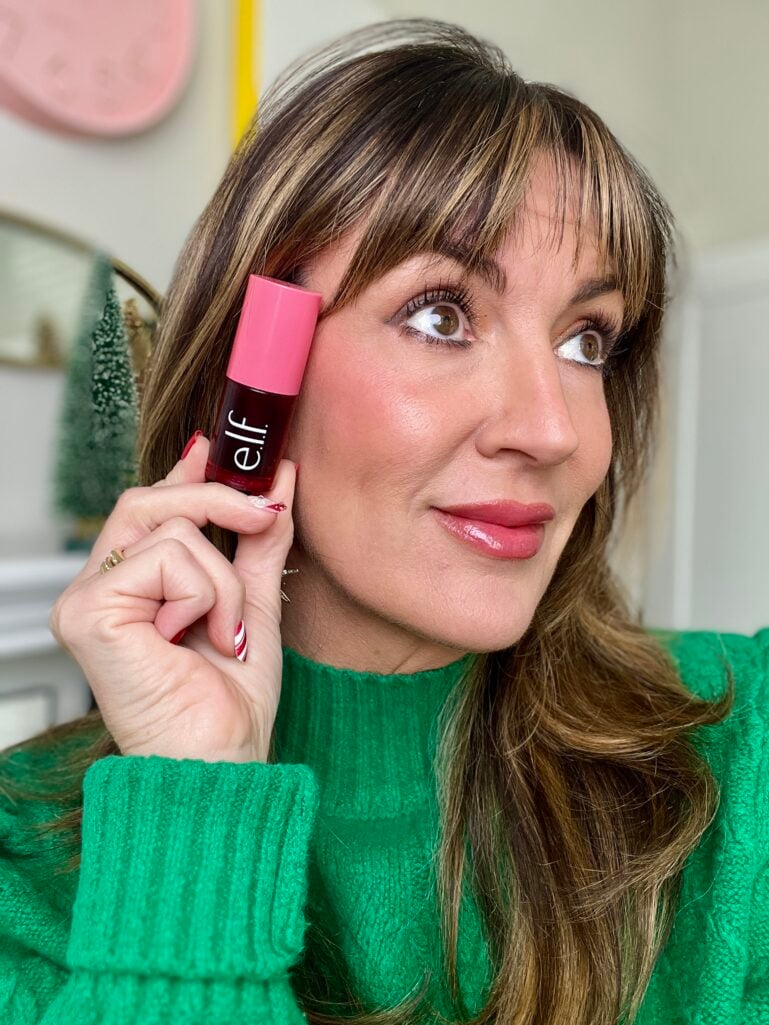 Woman holding e.l.f. Sheer For It cheek and lip stain in red shade near face, wearing green turtleneck with natural rosy cheek flush, mini Christmas trees visible in background