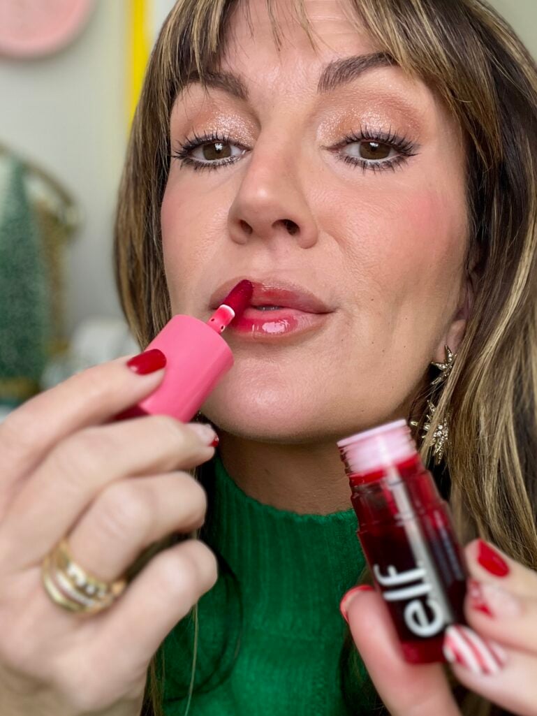Close-up of woman applying e.l.f. lip and cheek stain to lips with doe-foot applicator, showing shimmery champagne eyeshadow and glossy lip application technique