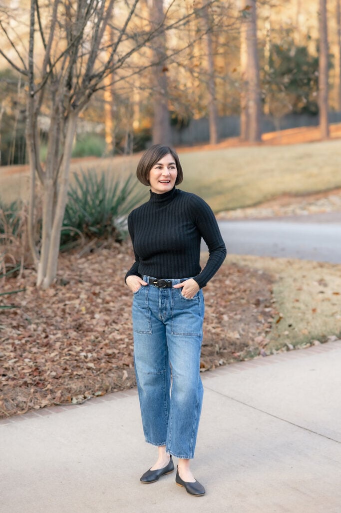 Kelly wearing petite barrel leg jeans with black ribbed turtleneck, black leather belt, and black ballet flats - full length front view showing curved silhouette of barrel leg style