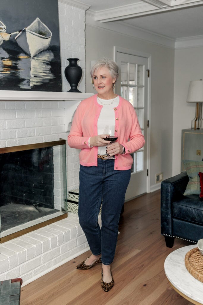 Woman over 50 wearing dark wash slim stretch jeans with coral pink cardigan and cream top, accessorized with leopard print belt and matching leopard flats, holding glass of wine by fireplace