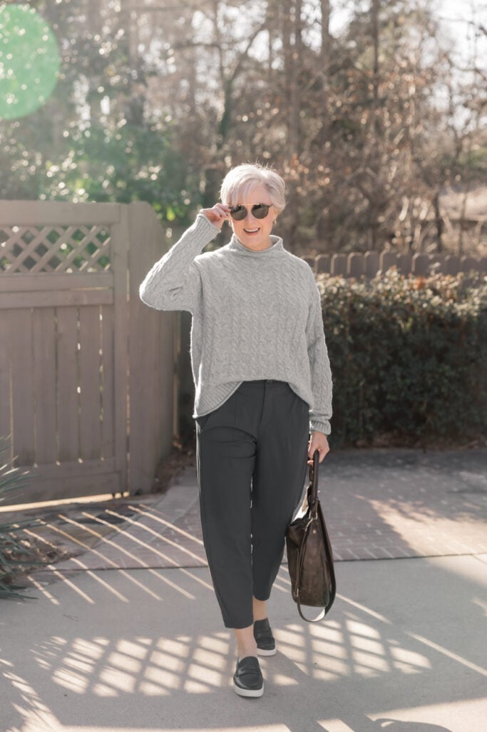 Woman over 50 wearing black tapered ankle pants with gray cable-knit turtleneck sweater and black slip-on sneakers, carrying a black leather tote bag