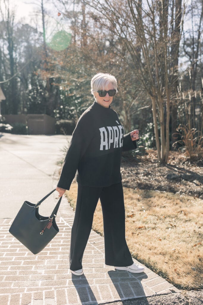 Woman over 50 wearing black wide-leg Cloud Plush pants with APRÈS sweater, styled with white sneakers and black Coach tote bag for cozy chic look