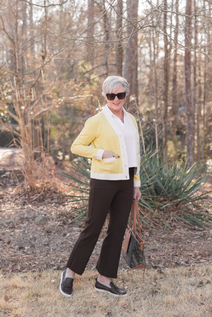 Woman over 50 wearing chocolate brown pull-on compression pants with yellow cardigan and white button-down shirt, styled with black loafers and camo tote bag