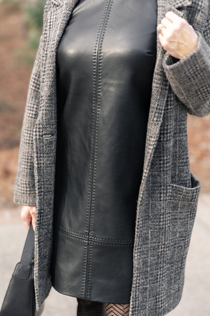 Detail shot of black faux leather mini dress showing seaming details and smooth finish layered under grey plaid coatigan with herringbone tights