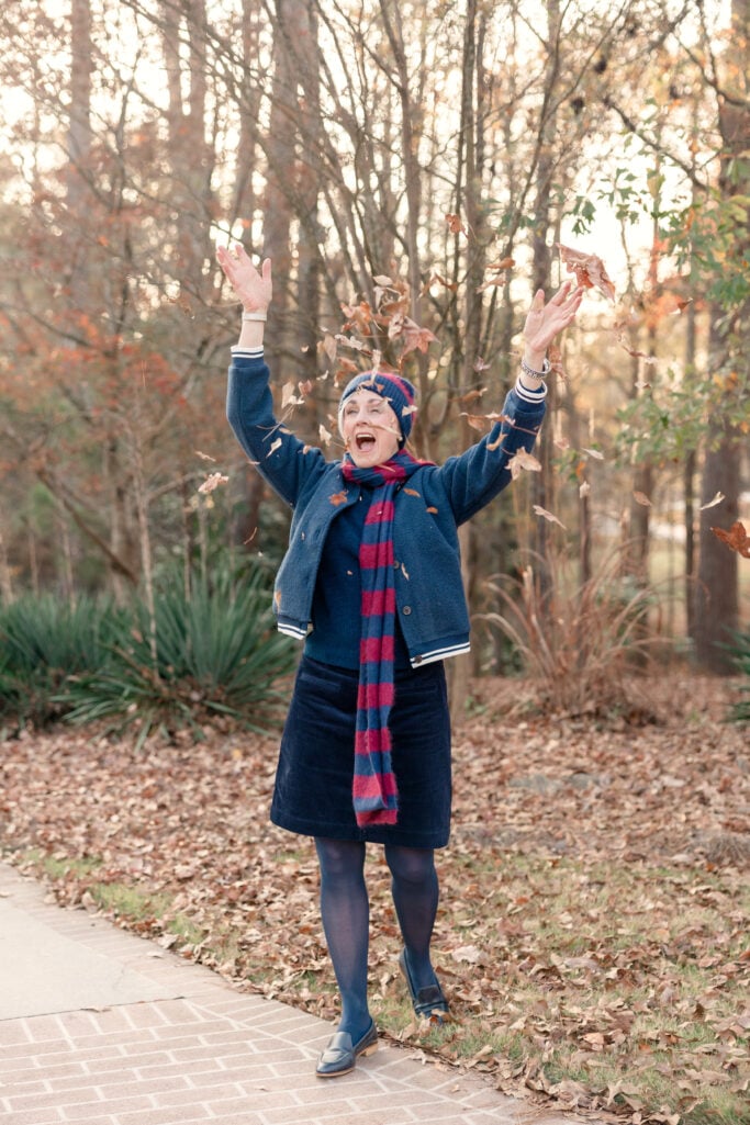 Beth Djalali in navy varsity-inspired sweater coat with cream collegiate striping navy corduroy skirt striped scarf and matching beanie tossing fall leaves