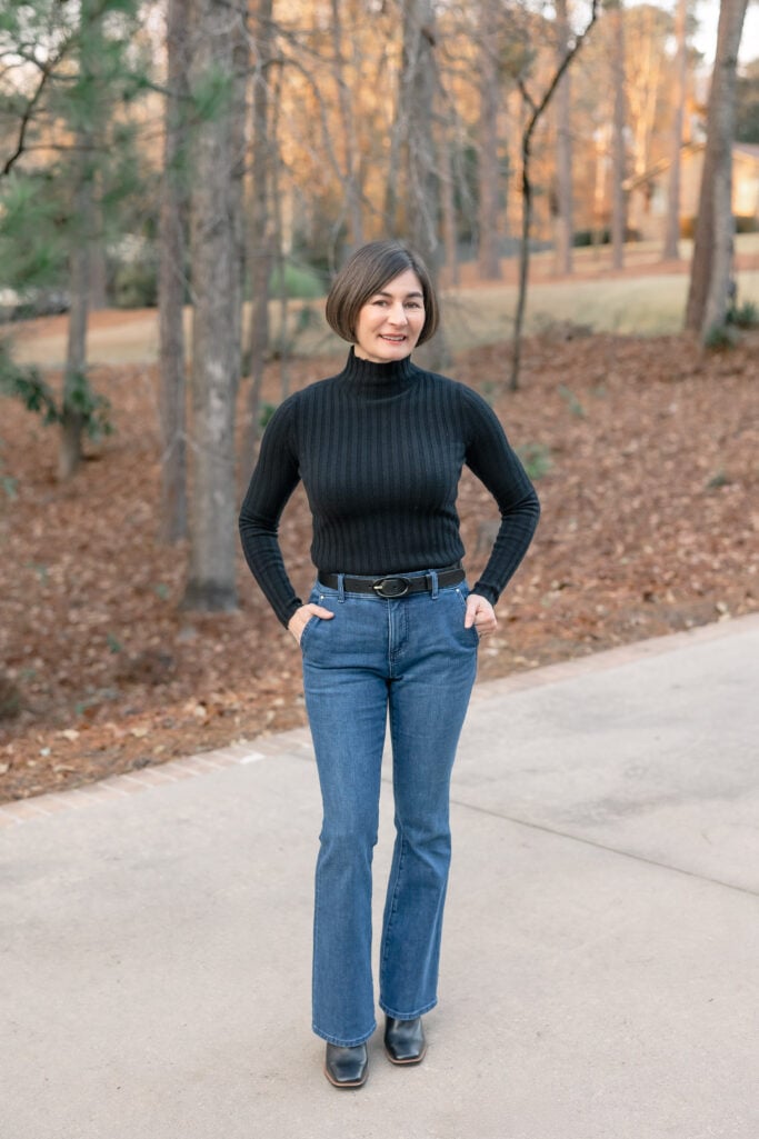 Kelly wearing petite bootcut jeans with black ribbed turtleneck, black belt, and black heeled boots - full length front view showing leg-lengthening bootcut silhouette
