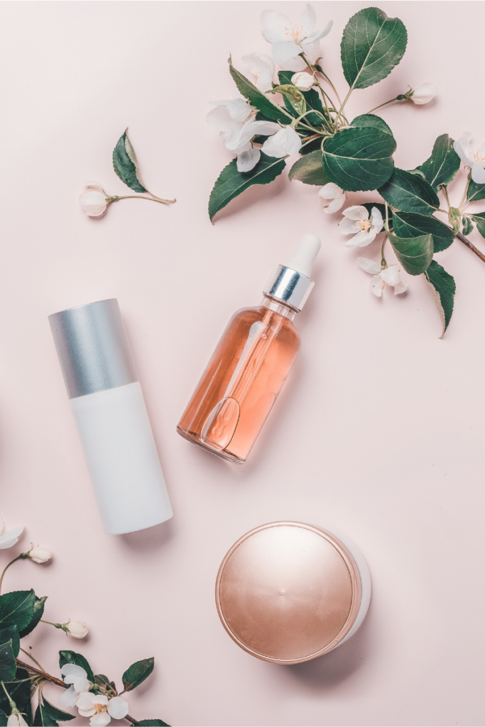 Flat lay of skincare products on a soft blush background, including a glass dropper serum bottle, minimalist lotion bottle, and round cream jar, styled with white blossoms and green leaves for a fresh, calming beauty scene.