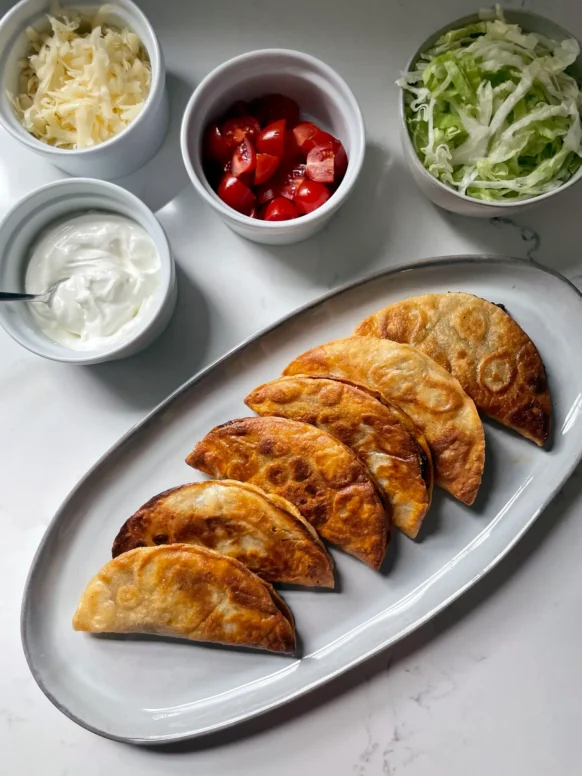 Six crispy tacos dorados on an oval platter with small bowls of shredded cheese, tomatoes, lettuce, and sour cream