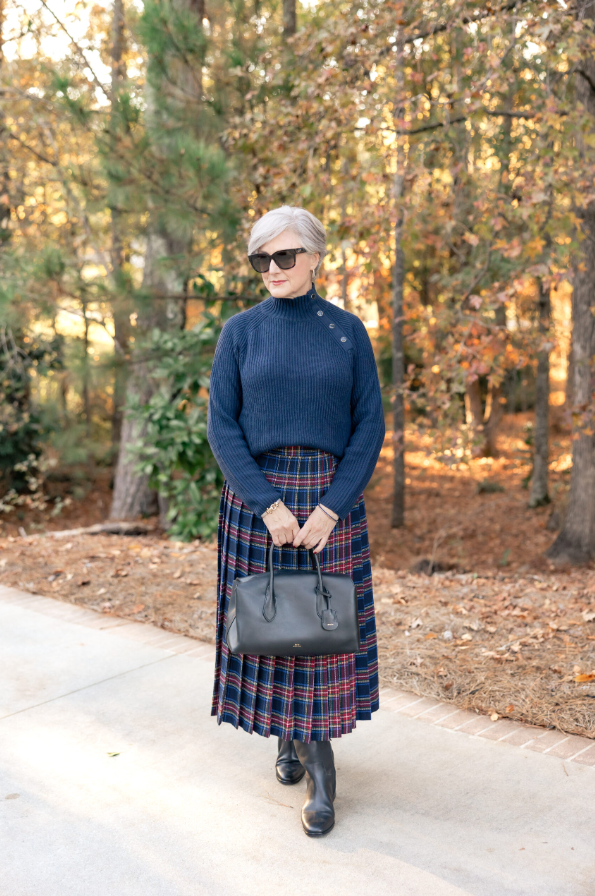 oman wearing a navy ribbed sweater tucked into a plaid pleated midi skirt, styled with black leather ankle boots and a structured black handbag, standing on a wooded walkway in autumn.