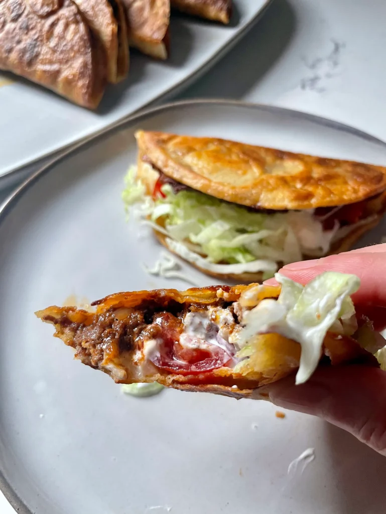 Hand holding a bitten crispy taco showing the beef filling, melted cheese, sour cream, and toppings inside